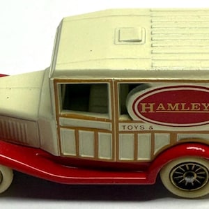 May include: A vintage toy truck with a cream-colored cab and roof, red fenders, and black wheels. The side of the truck features a Hamleys Toys & Games logo in a red oval.