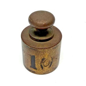 May include: A cylindrical brass weight with a dark, weathered patina. The top features a rounded handle knob. The side is marked with the black painted number 10, and the letters LB are stamped into the metal near the top edge of the weight.