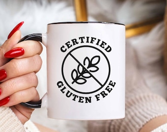 May include: A white ceramic mug with a black rim and handle, featuring the text "CERTIFIED GLUTEN FREE" in a black circular design. The mug is held by a person with red nail polish. The mug is part of a kitchenware collection.