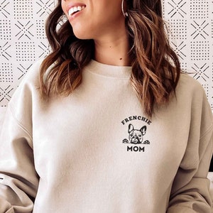 May include: A woman wearing a light beige sweatshirt with the text "Frenchie Mom" and a black graphic of a French bulldog peeking out from behind the text. She is sitting on a couch with a white throw blanket.