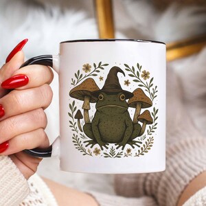 May include: White ceramic mug with a black rim and handle. The mug features a whimsical illustration of a green frog wearing a witch's hat, surrounded by mushrooms and foliage. The design is set against a white background.