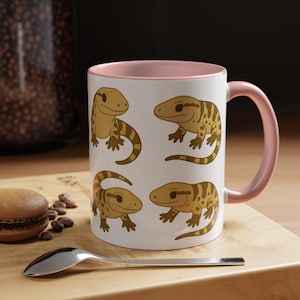 May include: White ceramic mug with a pink interior and handle. The mug features four cartoon geckos in shades of brown and yellow. A spoon and a macaron are on a wooden surface next to the mug.