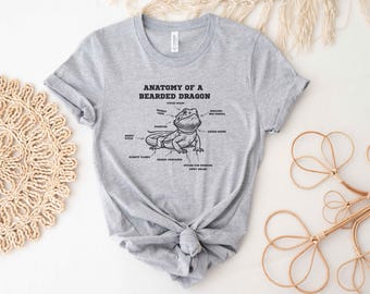 May include: A light gray t-shirt featuring a detailed black and white illustration of a bearded dragon with labeled anatomical parts. The text "ANATOMY OF A BEARDED DRAGON" is printed above the illustration. The shirt is tied in a knot at the bottom.