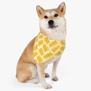May include: A tan Shiba Inu dog wearing a yellow bandana with a pattern of cheese slices. The bandana has a light yellow background.