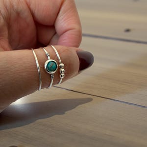 May include: A hand displaying four silver rings. One ring has a turquoise stone, another has three small beads. The rings are slender and delicate, demonstrating a minimalist jewellery style.
