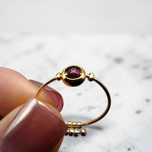 May include: A gold-colored ring with a round, dark red gemstone at the center. The ring has a delicate, adjustable band with small, decorative beads. The gemstone is set in a gold bezel, and the ring is held by a hand.