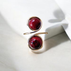 May include: A gold wire ring with two red gemstone beads. The ring is made of wire that is twisted and wrapped around the beads.