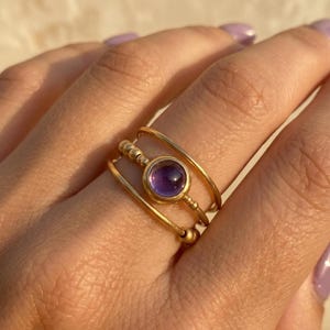 May include: A gold ring with a round, purple gemstone set in the center. The ring has a triple-band design, with a central band holding the gemstone and two outer bands. Small gold beads accent the ring's design.