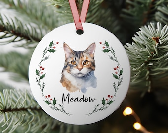 May include: White round Christmas ornament featuring a watercolor portrait of a cat with brown and orange fur. The cat is surrounded by a green holly wreath with red berries. The name "Meadow" is written in black script below the cat.