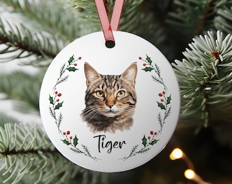 May include: A white, round Christmas ornament featuring a tabby cat portrait with the name "Tiger" in script. The cat is surrounded by a green and red holly wreath, and a red ribbon is attached for hanging.