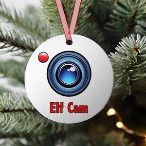 Könnte beinhalten: Weißer, runder Weihnachtsschmuck mit einem Kameralinsen-Design und dem roten Schriftzug "Elf Cam". Er hat ein rotes Band zum Aufhängen und einen roten Punkt. Der Schmuck hängt an einem Weihnachtsbaum.