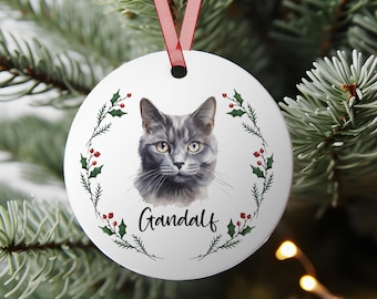 May include: A white, round Christmas ornament featuring a watercolor portrait of a gray cat with yellow eyes. The cat's name, "Gandalf," is written in a script font below the portrait. The ornament is encircled by a green and red holly wreath and has a red ribbon for hanging.