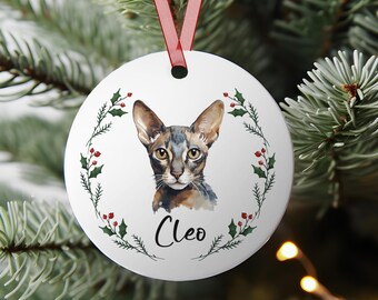 May include: A white, round Christmas ornament featuring a watercolor portrait of a cat named Cleo. The cat has brown and gray fur and is surrounded by a wreath of green leaves and red berries. A red ribbon is attached for hanging.