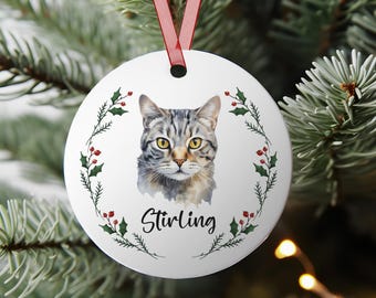May include: A white, round Christmas ornament featuring a watercolor portrait of a gray tabby cat with yellow eyes. The cat's name, "Stirling," is written in script below the image. A wreath of green holly and red berries encircles the cat.