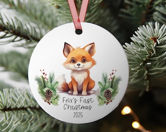 May include: Round white Christmas bauble featuring a cartoon fox in orange and white. The bauble has the text "Felix's First Christmas 2025" and is decorated with greenery and pine cones. A red ribbon is attached for hanging on a Christmas tree.