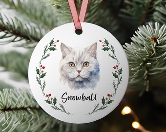 May include: A white ceramic Christmas ornament featuring a watercolor portrait of a gray cat with yellow eyes. The cat is surrounded by a green and red holly wreath. The name "Snowball" is written below the cat in a script font. A red ribbon is attached for hanging.