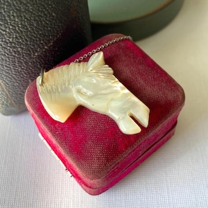May include: A pendant necklace featuring a carved horse head made of iridescent material, resting on a small, red velvet jewelry box. The horse head is attached to a delicate silver chain. The box is rectangular and has a hinged lid.