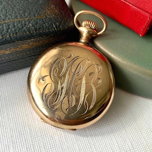 Puede incluir: Un reloj de bolsillo dorado con un monograma grabado. El reloj tiene una esfera circular y una tija de cuerda con un lazo. Se encuentra sobre un fondo de una caja verde y roja.