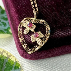 May include: A gold pendant necklace featuring a heart-shaped frame with a delicate filigree border. Inside the frame are two textured gold leaves, each accented with a small, round, vibrant pink gemstone. The pendant hangs from a thin gold chain.