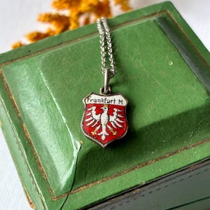 May include: A silver pendant necklace featuring a shield-shaped charm with red enamel and a white eagle emblem. The charm displays the text Frankfurt M at the top. The pendant hangs from a delicate silver chain, showcasing a detailed heraldic design.