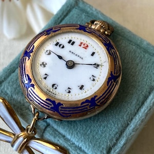 May include: A gold-tone pocket watch with a white face, black numerals, and a red number 12. The watch features a blue enamel decorative border and is attached to a white and gold-tone bow-shaped pin. The word Bristol is printed on the dial.