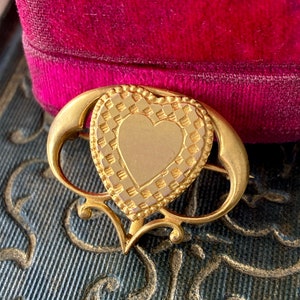 Puede incluir: Un broche de corazón de oro con un patrón de cuadros alrededor del borde. El corazón tiene una superficie lisa y en blanco en el centro.