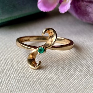 Pode incluir: Um anel dourado com um design curvo único. O anel apresenta uma pequena pedra preciosa verde esmeralda redonda no centro do design. O anel está sobre um fundo macio e neutro, com toques de flores roxas ao fundo.