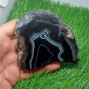 K&ouml;nnte beinhalten: Ein polierter, schwarzer Achatstein mit wei&szlig;en und hellblauen B&auml;ndern. Der Naturstein mit rauen Kanten wird in einer Hand gehalten. Das Muster des Achats ist eine beeindruckende Darstellung nat&uuml;rlicher Kunstfertigkeit.