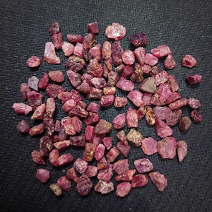 May include: A scattering of small, rough-cut ruby gemstones. The stones vary in size and shape, displaying a range of red and pink hues. The gemstones are scattered on a dark surface, creating a contrast that highlights their colour and texture.