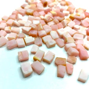 Op de afbeelding: Een verzameling kleine, onregelmatig gevormde, roze en witte edelstenen. De edelstenen zijn verspreid over een wit oppervlak, met verschillende roze tinten en sommige met een doorschijnende kwaliteit. Het algemene effect is een zacht, pastelkleurenpalet.