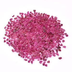 May include: A pile of small, rough-cut ruby gemstones. The stones are a deep, rich pinkish-red color, with variations in tone and translucency. The gemstones are scattered across a white surface.