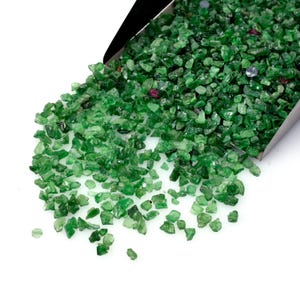 May include: A metal scoop filled with small, raw, green gemstone fragments. The pile of stones features various shades of translucent and opaque green, with a few scattered small red and clear crystalline pieces mixed throughout the collection.