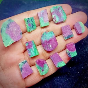 以下が含まれることがあります： ピンクとグリーンの色合いが混ざった、原石のルビーの結晶のコレクション。宝石は不規則な形をしており、サイズも様々で、自然な質感と模様が際立っています。暗い背景の中で、手の中に結晶が収められています。