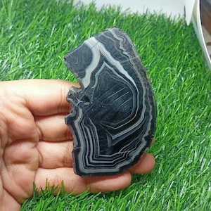 K&ouml;nnte beinhalten: Ein polierter Achatstein mit unregelm&auml;&szlig;iger Form und einem auff&auml;lligen Muster aus schwarzen, wei&szlig;en und grauen B&auml;ndern. Der Stein wird in einer Hand gehalten, wodurch sein nat&uuml;rliches, geschichtetes Design zur Geltung kommt. Die Oberfl&auml;che des Achats ist glatt und reflektierend.