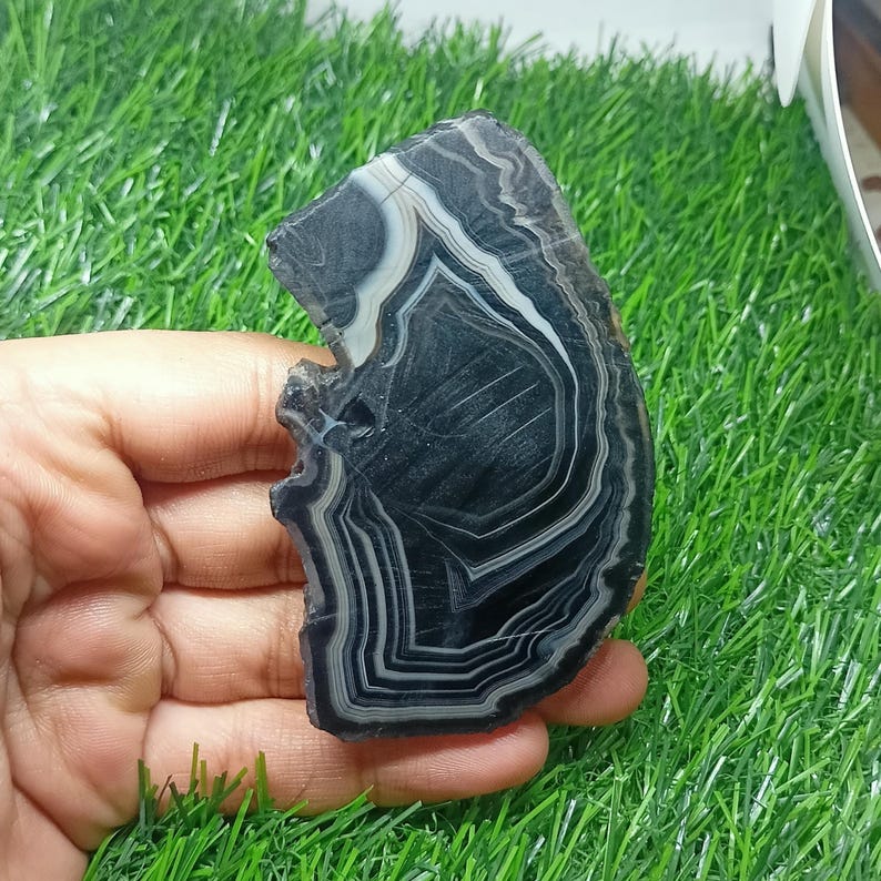 K&ouml;nnte beinhalten: Ein polierter Achatstein mit unregelm&auml;&szlig;iger Form und einem auff&auml;lligen Muster aus schwarzen, wei&szlig;en und grauen B&auml;ndern. Der Stein wird in einer Hand gehalten, wodurch sein nat&uuml;rliches, geschichtetes Design zur Geltung kommt. Die Oberfl&auml;che des Achats ist glatt und reflektierend.