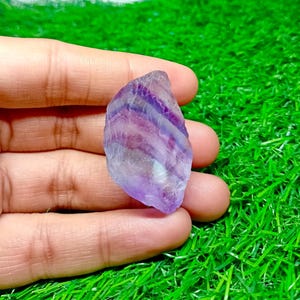 Puede incluir: Cristal de fluorita en bruto, con tonos morados y blancos. La piedra tiene una apariencia en capas y una textura rugosa e irregular. El cristal se sostiene sobre un fondo verde.