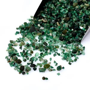 Può includere: Una collezione di piccole pietre preziose di smeraldo grezze in varie tonalità di verde, versate da una paletta di metallo su una superficie bianca. Le pietre preziose variano per dimensioni e forma, alcune appaiono traslucide.