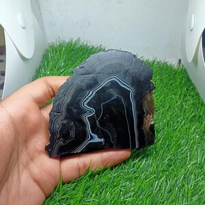 K&ouml;nnte beinhalten: Eine polierte, schwarze Achatscheibe mit wei&szlig;en und hellblauen B&auml;ndern. Der Naturstein hat einen rauen, unebenen Rand. Der Stein wird in einer Hand gehalten.