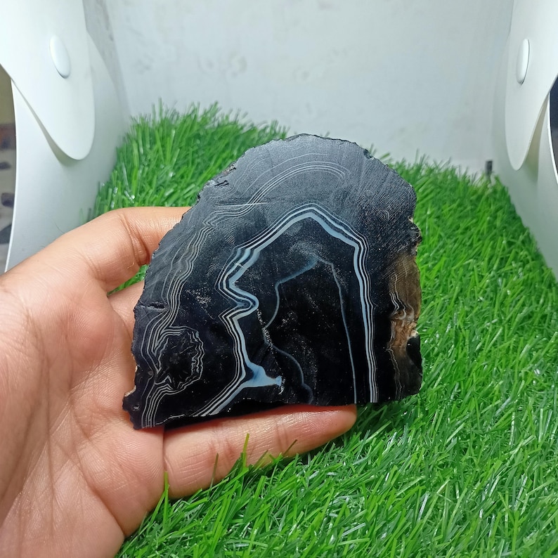 K&ouml;nnte beinhalten: Eine polierte, schwarze Achatscheibe mit wei&szlig;en und hellblauen B&auml;ndern. Der Naturstein hat einen rauen, unebenen Rand. Der Stein wird in einer Hand gehalten.