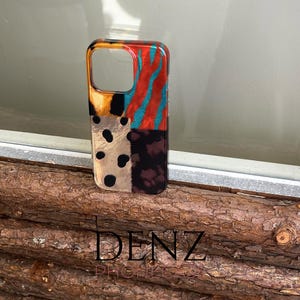 May include: A patterned phone case in a vibrant design. The case combines animal print, stripes, and polka dots in orange, teal, black, and brown. The brand name "DENZ PHONES" is printed at the bottom. A stylish accessory for a mobile phone.