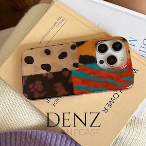 May include: A patterned phone case with a clear border. The case has sections with leopard print, a dark maroon pattern, orange and teal stripes. The text "DENZ" and "ONECASE" are visible on the book below the phone case.