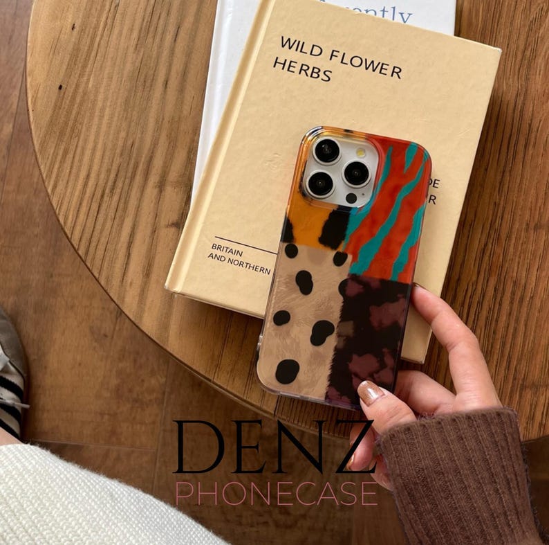 May include: A patterned phone case in shades of orange, teal, brown, and black, held over a book titled "Wild Flower Herbs" and another book with the text "Britain and Northern". The phone case has a patchwork design. The brand name "DENZ PHONECASE" is visible.