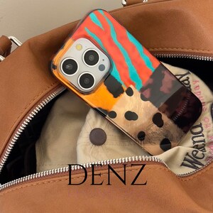 May include: A patterned phone case in shades of orange, teal, and brown, resting inside a brown leather bag. The case has a clear border and multiple camera lenses. The bag's zipper is visible, along with the text "DENZ".