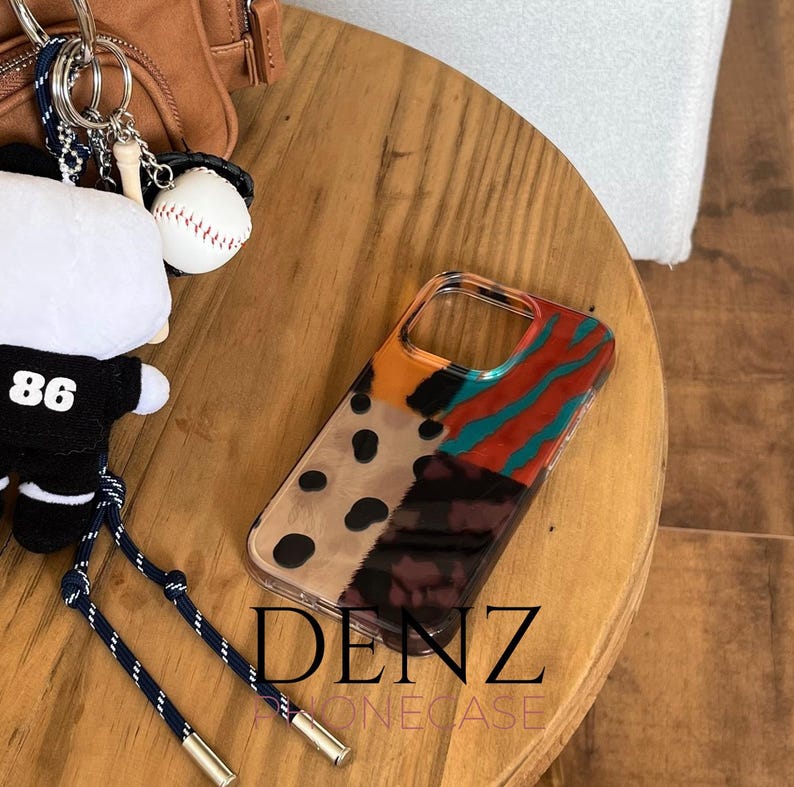 May include: A clear phone case with a vibrant abstract design. The design features sections of orange, teal, red, black spots, and a leopard print pattern. The text "DENZ PHONECASE" is printed at the bottom of the case.
