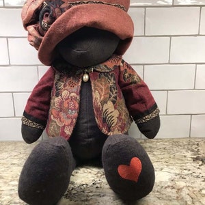 Op de afbeelding: Een zwarte stoffen pop die een rood en bruin bloemenpatroon jasje draagt en een rode hoed met een stoffen roos. De pop heeft een hartvormige patch op één voet.