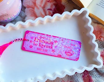 Puede incluir: Un marcapáginas rosa y morado con el texto "Friends to Lovers Book Club" y una borla rosa. El marcapáginas está sobre una bandeja blanca con borde festoneado, junto a un libro y una vela.