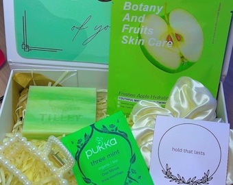 May include: A gift box with self-care items. Includes a green face mask, a bar of soap, a tea bag, a pearl necklace, a bath bomb, and chocolates. The box has the words "Take care of you" on the lid.