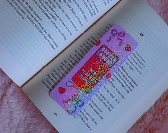 Puede incluir: Un marcapáginas colorido con acabado holográfico, con un diseño de caja de cerillas con el texto "The Perfect Match". El marcapáginas está colocado en un libro abierto.