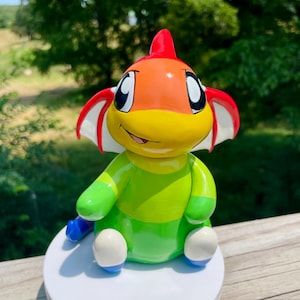 May include: A colorful, hand-painted ceramic figurine of a cartoon-like creature. The figurine features a red fin, an orange and yellow head, and a green body. It has white and blue accents and is sitting on a white base.