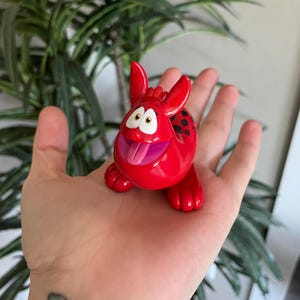 Op de afbeelding: Een felrode cartoonfiguur met grote oren, een lachend gezicht en een uitstekende tong. De figuur heeft zwarte vlekken op de rug en rust op een hand.